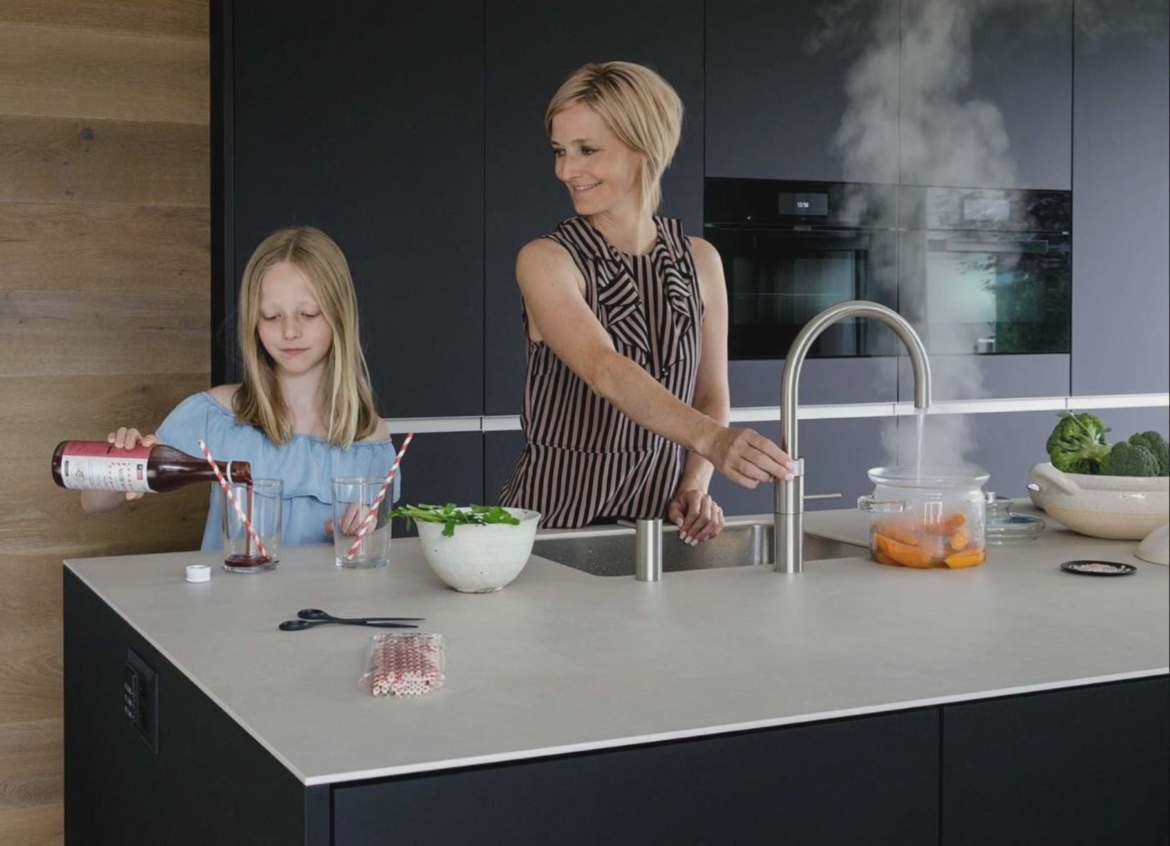 Charger la vidéo: Vrouw schakelt de Quooker kraan aan in een luxe keuken en haar kind scheent rode wijn in.