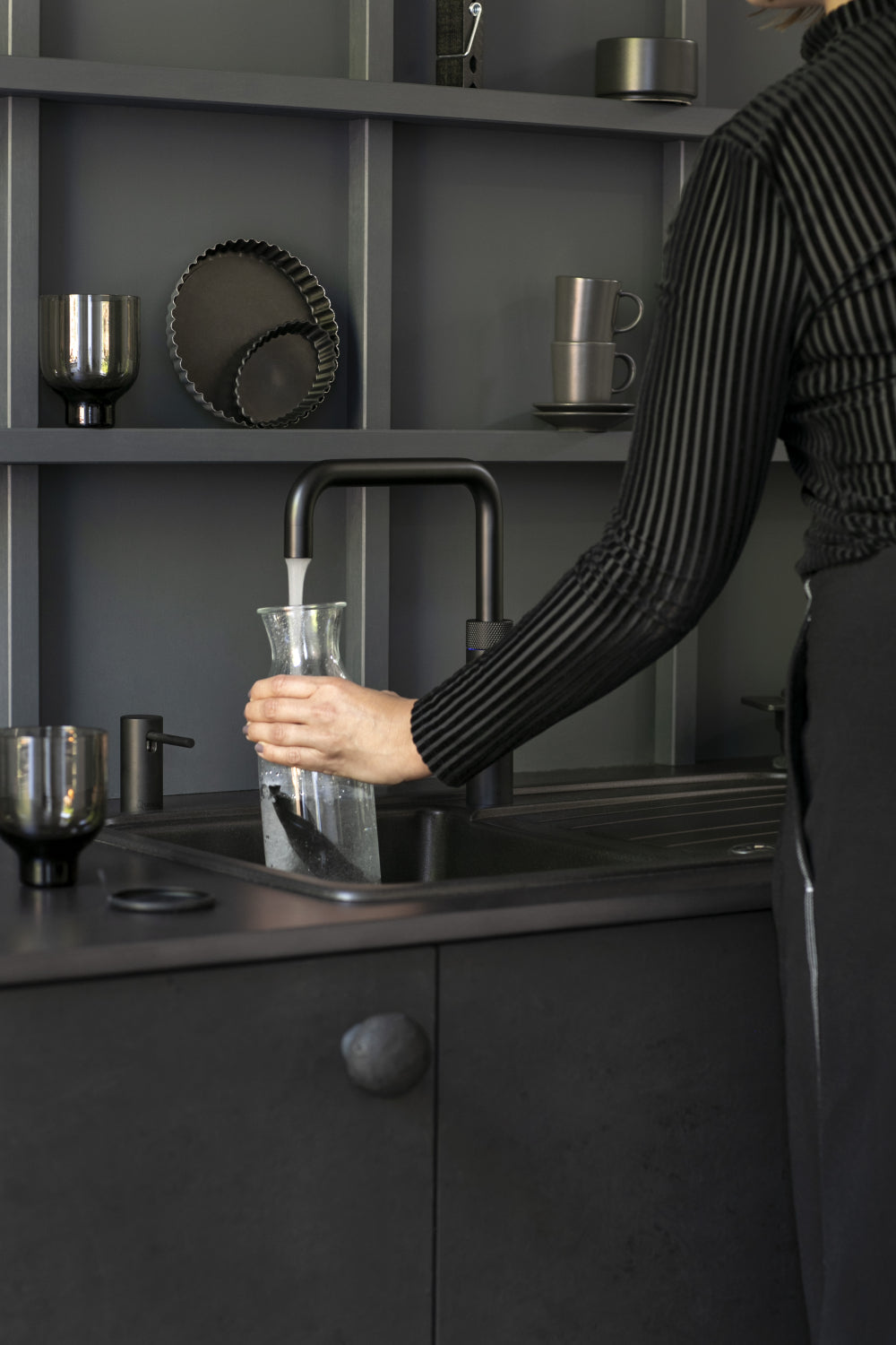 Persoon vult glazen karaf met water uit een zwarte Quooker kokendwaterkraan in een moderne donkere keuken
