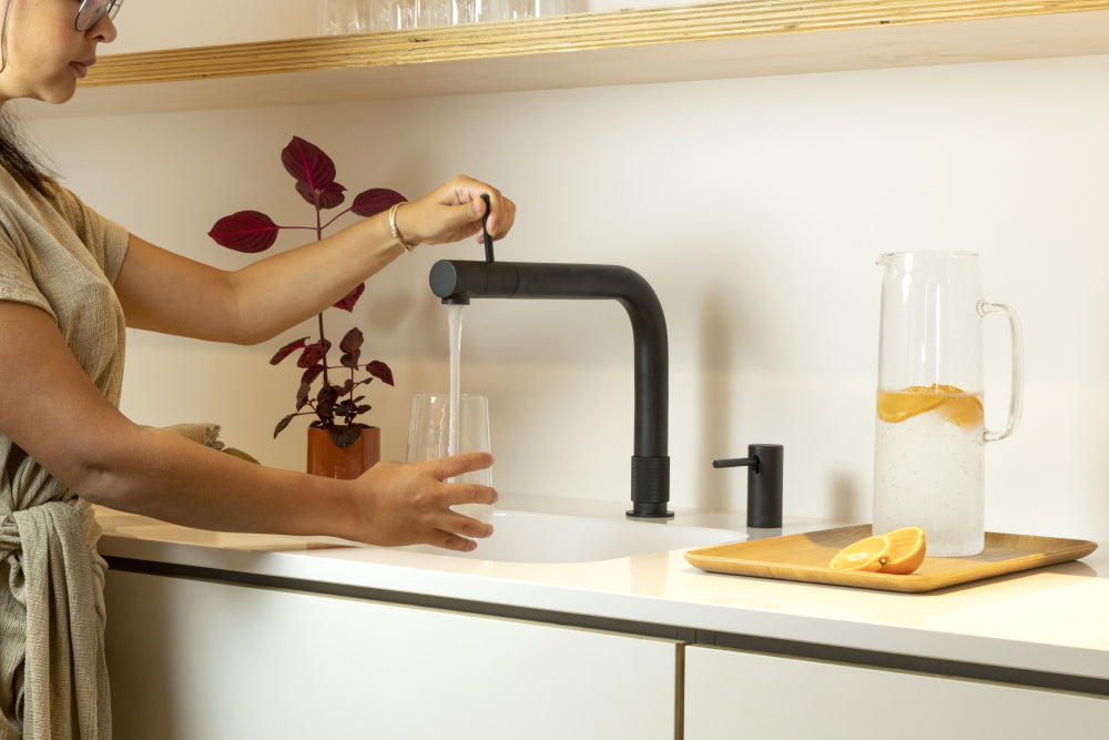 Vrouw vult glas met water uit een moderne zwarte Quooker kokendwaterkraan in lichte keuken met karaf citruswater